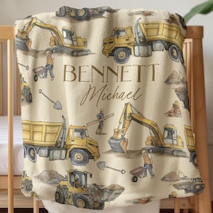 Könnte beinhalten: Eine beige Babydecke mit Illustrationen von gelben Baumaschinen, Arbeitern, Schaufeln und den Namen "BENNETT Michael" in brauner Schrift. Die Decke ist über ein Holzbettchen drapiert.