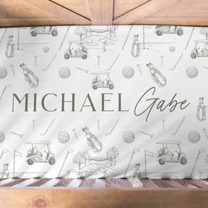 K&ouml;nnte beinhalten: Ein wei&szlig;es Kinderbettlaken mit einem Golf-Themenmuster. Das Design umfasst Golfwagen, Golfb&auml;lle, Golftaschen und Golfschl&auml;ger in hellgr&uuml;ner Farbe. Der Name "MICHAEL Gabe" ist in einer passenden gr&uuml;nen Schrift gedruckt.