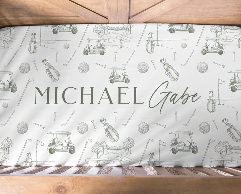 K&ouml;nnte beinhalten: Ein wei&szlig;es Kinderbettlaken mit einem Golf-Themenmuster. Das Design umfasst Golfwagen, Golfb&auml;lle, Golftaschen und Golfschl&auml;ger in hellgr&uuml;ner Farbe. Der Name "MICHAEL Gabe" ist in einer passenden gr&uuml;nen Schrift gedruckt.
