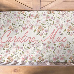 May include: A crib sheet with a floral pattern in pink, peach, and green. The sheet features the name "Caroline Mae" in a cursive, pink font. The design includes flowers and bows on a white background.