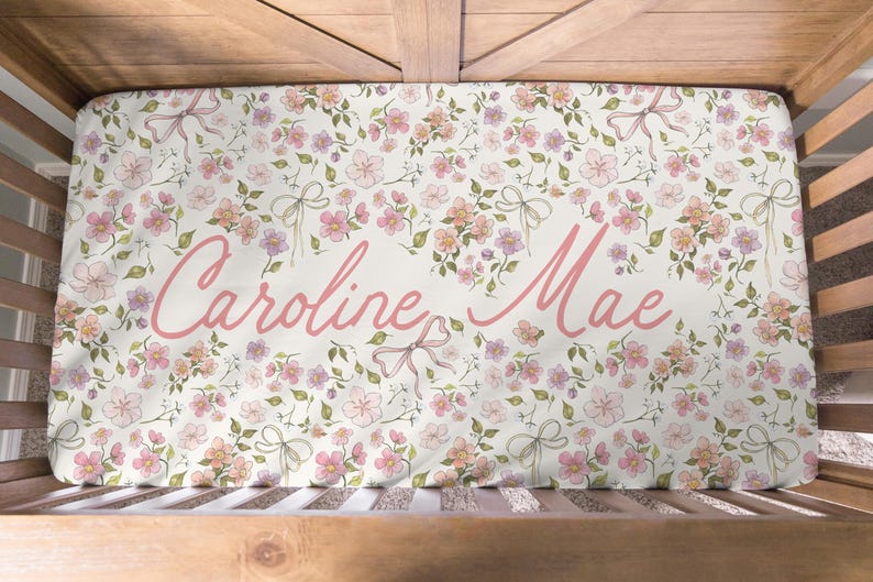 May include: A crib sheet with a floral pattern in pink, peach, and green. The sheet features the name "Caroline Mae" in a cursive, pink font. The design includes flowers and bows on a white background.