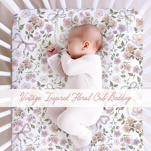 May include: A baby sleeps peacefully in a white crib on a floral patterned crib sheet. The sheet features pink, purple, and peach flowers with green leaves and pink bows on a white background. Text on the image reads "Vintage Inspired Floral Crib Bedding."