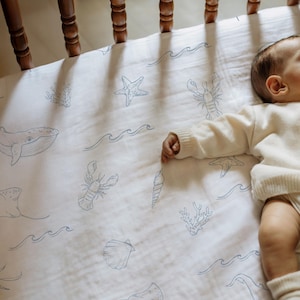 May include: A white baby crib sheet with a blue sea life pattern, including whales, starfish, and seashells. The sheet is made of soft fabric and is designed for a crib. The sheet is part of a nursery collection.