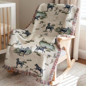 May include: A cream-colored woven throw blanket with a black labrador retriever dog pattern. The blanket is draped over a wooden rocking chair.