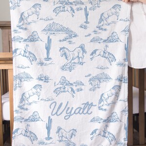 Puede incluir: Manta blanca con un patr&oacute;n azul de caballos y paisaje des&eacute;rtico. La manta presenta ilustraciones de caballos, jinetes, monta&ntilde;as y cactus. El nombre "Wyatt" est&aacute; escrito en azul.