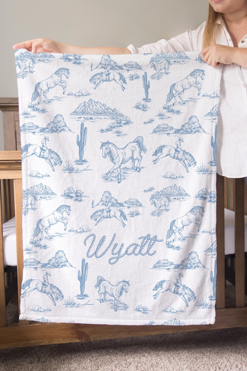 Puede incluir: Manta blanca con un patr&oacute;n azul de caballos y paisaje des&eacute;rtico. La manta presenta ilustraciones de caballos, jinetes, monta&ntilde;as y cactus. El nombre "Wyatt" est&aacute; escrito en azul.
