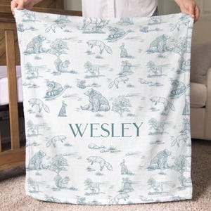 May include: A white baby blanket with a blue toile pattern featuring bears, foxes, raccoons, and rabbits. The name "WESLEY" is printed in blue block letters across the center of the blanket. The blanket is held up by a person.