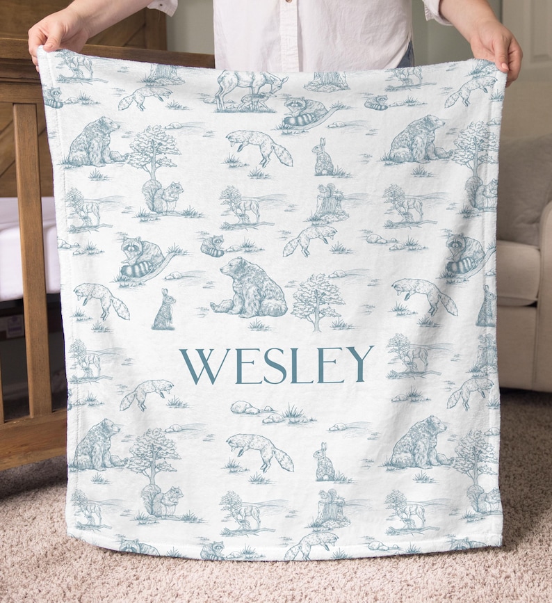 May include: A white baby blanket with a blue toile pattern featuring bears, foxes, raccoons, and rabbits. The name "WESLEY" is printed in blue block letters across the center of the blanket. The blanket is held up by a person.