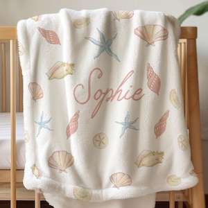 May include: A cream-coloured baby blanket adorned with a seashell and starfish design. The blanket showcases seashells in pink, peach, and yellow, alongside blue starfish. The name "Sophie" is written in pink script across the blanket.