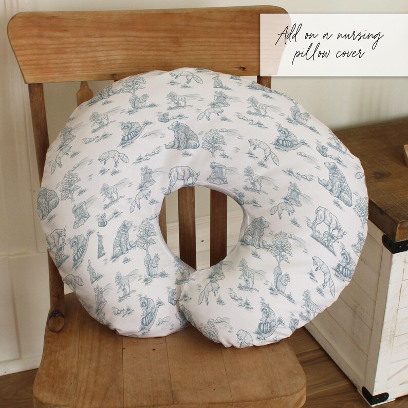 May include: A white nursing pillow cover with a blue toile pattern featuring bears, foxes, and trees. The pillow is circular with a crescent-shaped cutout. The pillow is on a wooden chair.