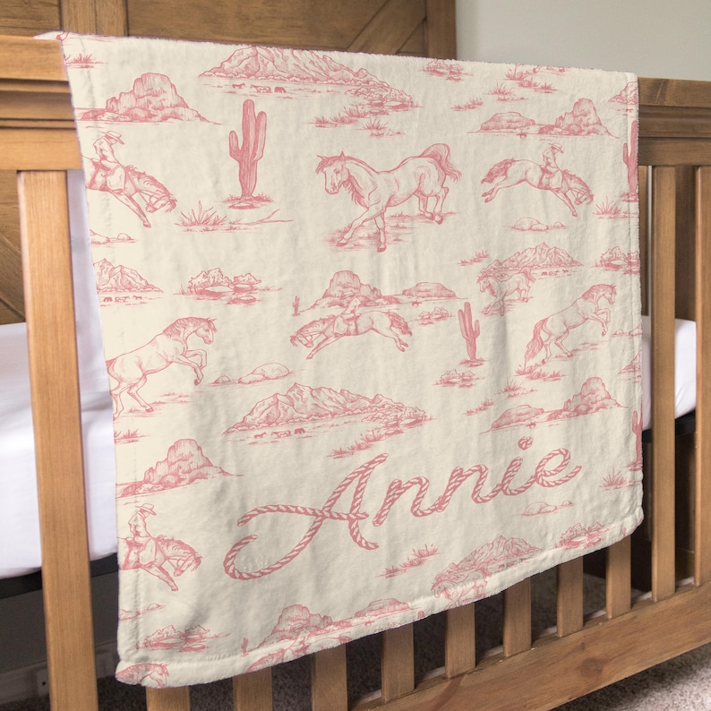 Puede incluir: Una manta para beb&eacute; de color crema con un estampado de toile rosa con tem&aacute;tica del oeste. El dise&ntilde;o incluye vaqueros, caballos, cactus y monta&ntilde;as. El nombre "Annie" est&aacute; escrito en una fuente de estilo cuerda.