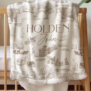 May include: A cream-colored baby blanket with a farm scene design in brown tones. The blanket features illustrations of farm animals, trees, and fences. The name "Holden John" is printed in a decorative font.