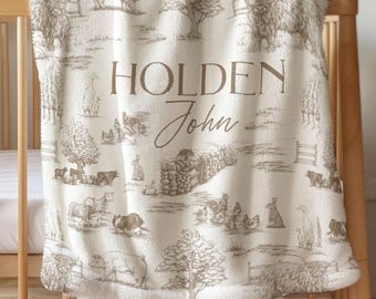 May include: A cream-colored baby blanket with a farm scene design in brown tones. The blanket features illustrations of farm animals, trees, and fences. The name "Holden John" is printed in a decorative font.