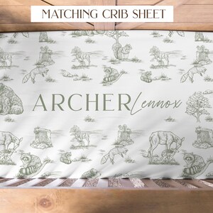 May include: White crib sheet with a woodland animal pattern in green. The design includes bears, deer, foxes, raccoons, and squirrels. The name "Archer Lennox" is printed in green. The text "MATCHING CRIB SHEET" is at the top.