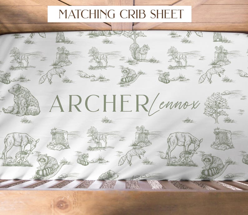 May include: White crib sheet with a woodland animal pattern in green. The design includes bears, deer, foxes, raccoons, and squirrels. The name "Archer Lennox" is printed in green. The text "MATCHING CRIB SHEET" is at the top.