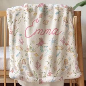 Peut inclure: Une couverture de bébé crème avec un motif de sirène et de vie marine. La couverture présente des sirènes, des coquillages, des étoiles de mer et des poissons aux couleurs pastel. Le nom "Emma" est écrit en rose sur la couverture.