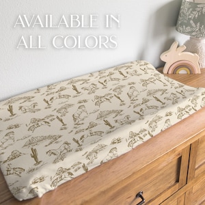 Puede incluir: Un cambiador beige con un estampado marr&oacute;n de caballos, cactus y monta&ntilde;as. El texto blanco dice "AVAILABLE IN ALL COLORS". El cambiador est&aacute; sobre una c&oacute;moda de madera.