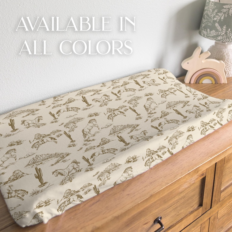 Puede incluir: Un cambiador beige con un estampado marr&oacute;n de caballos, cactus y monta&ntilde;as. El texto blanco dice "AVAILABLE IN ALL COLORS". El cambiador est&aacute; sobre una c&oacute;moda de madera.