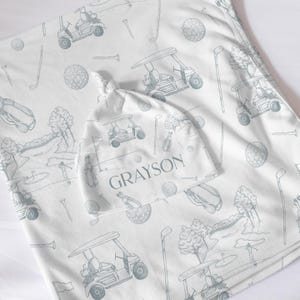May include: A white baby blanket and matching hat set with a golf-themed pattern in light blue. The pattern includes golf carts, golf balls, and golf clubs. The hat is personalized with the name "Grayson".