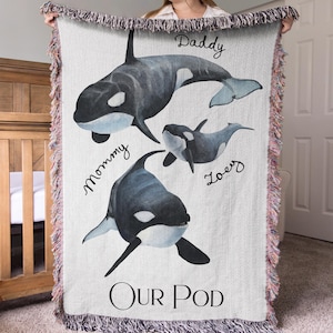 May include: A woven throw blanket featuring a watercolor illustration of three orca whales. The text Daddy, Mommy, Zoey, and Our Pod is printed on the white fabric. The blanket has a multicolored fringed edge on all four sides of the rectangular textile.