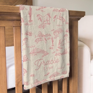 Puede incluir: Una manta para beb&eacute; de color crema con un estampado repetido de caballos, cactus y monta&ntilde;as en un tono rosa-rojo. La manta est&aacute; sobre una cuna de madera y presenta el nombre "Gracie Lynn" en una fuente cursiva.
