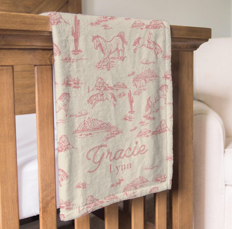Puede incluir: Una manta para beb&eacute; de color crema con un estampado repetido de caballos, cactus y monta&ntilde;as en un tono rosa-rojo. La manta est&aacute; sobre una cuna de madera y presenta el nombre "Gracie Lynn" en una fuente cursiva.