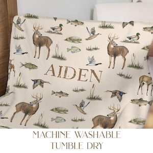 May include: A beige blanket featuring a repeating pattern of deer, ducks, fish, and reeds. The name "AIDEN" is printed in large brown letters. The blanket also has the words "MACHINE WASHABLE" and "TUMBLE DRY" printed on it.