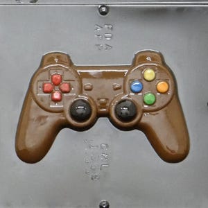 May include: A brown plastic mold for making chocolate game controllers. The mold has a detailed design of a game controller with buttons and joysticks. The mold is in a clear plastic case.