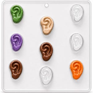 May include: A clear plastic rectangular mold featuring nine recessed cavities shaped like human ears. Several cavities are filled with colored substances in shades of green, tan, purple, dark brown, light brown, white, and orange, while others remain empty.