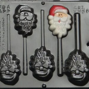 May include: A gray plastic mold for making candy lollipops. The mold has five cavities, four of which are shaped like Santa Claus faces and one is shaped like a snowman. The mold has a stick holder for each cavity.