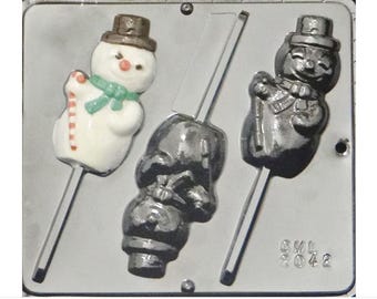 Puede incluir: Un molde de plástico gris para hacer piruletas de muñeco de nieve. El molde tiene dos formas de muñeco de nieve, una con un sombrero blanco y la otra con un sombrero negro. El molde también tiene dos palos para las piruletas.