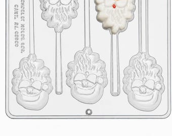 Könnte beinhalten: Eine transparente Kunststoff-Schokoladenform mit fünf Aussparungen in Form von Weihnachtsmann-Gesichtern und integrierten Stielkanälen. Eine Aussparung zeigt eine bemalte Weihnachtsmannfigur mit roter Mütze, weißem Bart und pfirsichfarbenem Gesicht.