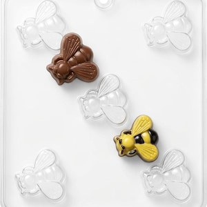 May include: A clear plastic rectangular candy mold featuring six recessed bee-shaped cavities. One cavity contains a solid brown chocolate bee, and another contains a yellow and black chocolate bee, while the remaining four cavities are empty.