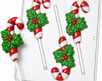 Peut inclure: Un moule à bonbons en plastique transparent présentant trois cavités en forme de sucettes canne de Noël avec des feuilles de houx et des baies. Une sucette finie aux rayures rouges et blanches avec des feuilles de houx est posée à gauche.