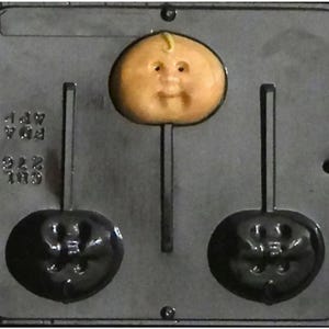 May include: A black plastic mold for making lollipops. The mold has two cavities, each shaped like a face. The mold also has a small, round cavity for making a lollipop head. The mold is designed to make two lollipops at a time.