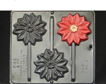 Puede incluir: Un molde de plástico negro y rojo para hacer piruletas. El molde tiene cuatro cavidades, cada una con forma de flor con un palo adjunto. El molde está etiquetado como "FDA Approved" y "2800 Z".