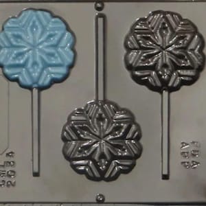 May include: A silver snowflake lollipop mold with three cavities. The mold has a blue snowflake in one cavity and two black snowflakes in the other cavities. The mold is labeled "FDA APB" and "CMLL 203A".