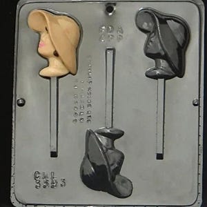 May include: A silver candy mold with two cavities for making chocolate lollipops. Each cavity features a woman's head wearing a hat. One cavity has a woman with a light brown hat and the other has a woman with a black hat.