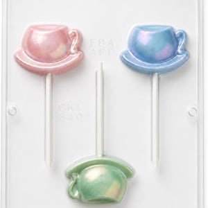 May include: A clear plastic candy mold containing three tea cup shaped lollipops with white sticks. The lollipops are iridescent pink, blue, and green. The mold features the embossed text FDA APP and CKL 3407 on the clear plastic surface.
