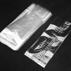 May include: A stack of clear cellophane bags, alongside a single bag. The bags are rectangular and appear to be empty. The image is in black and white, with the bags reflecting light.