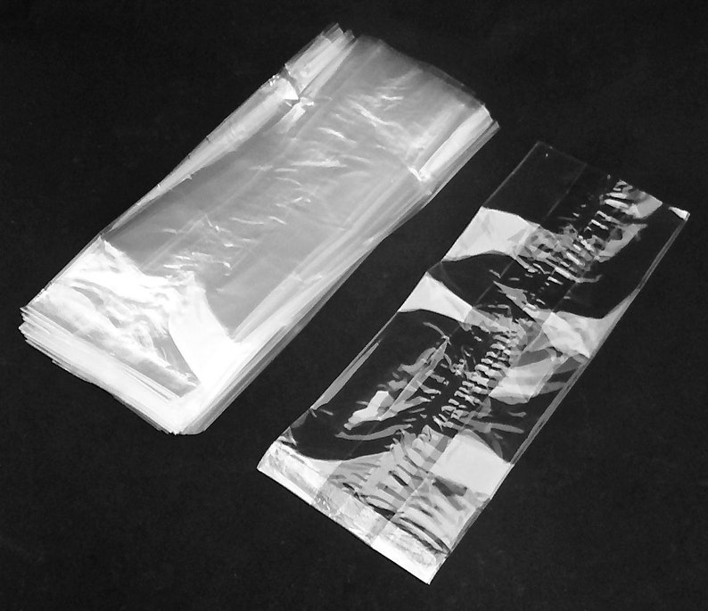 May include: A stack of clear cellophane bags, alongside a single bag. The bags are rectangular and appear to be empty. The image is in black and white, with the bags reflecting light.