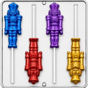 May include: A clear plastic candy mold featuring four nutcracker-shaped cavities. Two nutcrackers are oriented upright in blue and purple, while two are oriented upside down in red and gold. Each cavity includes a slot for a white lollipop stick.