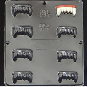 May include: A black plastic mold with eight cavities for making chocolate vampire teeth. The mold has the text "GML 847" and "FDA APP" on it.