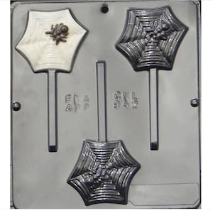May include: A silver candy mold with three spider web designs. Each design has a spider in the center. The mold has two stick holders. The mold is labeled "FDA APP" and "GWL 942".