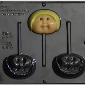May include: A gray plastic mold for making lollipops. The mold features a smiling doll face lollipop and two lollipop shapes. The doll face has yellow hair and a peach-colored face. The mold has text on it.