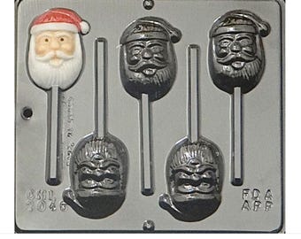 Puede incluir: Un molde de plástico gris para hacer piruletas de caramelo. El molde tiene cinco cavidades, cuatro de las cuales tienen forma de cabeza de Papá Noel y una tiene forma de cara genérica. El molde está etiquetado como "CML 2046" y "FDA APF".