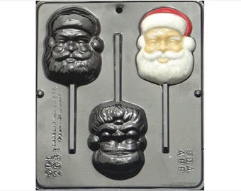 Puede incluir: Un molde de plástico gris para hacer piruletas de caramelo. El molde tiene tres cavidades, cada una con forma de cara. Una cavidad es una cara de Papá Noel con un gorro rojo, otra es una cara en blanco y negro con barba, y la tercera es una cara de monstruo en blanco y negro.