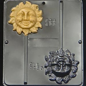 May include: A silver metal mold for making candy lollipops. The mold has two cavities, each shaped like a sun with a face. One sun is gold and the other is black.