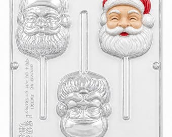 May include: A clear plastic candy mold featuring three Santa Claus head shapes with integrated stick channels. One Santa is painted with a red hat, white beard, and peach face, while the other two are transparent. Text reads OMI 203.
