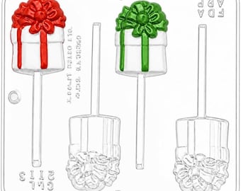 May include: A clear plastic candy mold featuring four cavities shaped like gift boxes with bows on sticks. Two cavities are filled with red and green paint, while two remain clear. The mold includes text reading FDA APP and 2113.